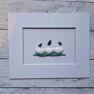 Peut inclure: Une œuvre encadrée représentant trois oiseaux perchés sur des coquillages. Les oiseaux sont bleu foncé et noirs, et les coquillages sont blancs. Les coquillages reposent sur une base turquoise, le tout dans un cadre blanc.
