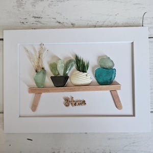 May include: A white framed wall art piece featuring a wooden shelf with four small potted plants made of sea glass and natural elements. The plants sit on the shelf, and the word Home is written in a wooden script font below the shelf on the white background.