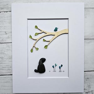 May include: Framed artwork depicting a black pebble dog under a wooden branch with a blue bird and green leaves. Small blue flowers are in the foreground. The artwork is set against a white background.