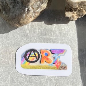 May include: A rectangular sticker with rounded corners featuring the word ART in colourful, hand-drawn lettering. The letter A is inside a black circle, the R is orange, and the T is formed by a blue lightning bolt above a small red heart.
