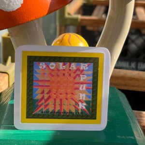 May include: A square sticker with a white border featuring a stylised orange sun graphic with radiating points. The text "SOLAR PUNK" is printed in white letters over the sun, and the text "artifactmuse.etsy.com" is printed in small black letters at the bottom.