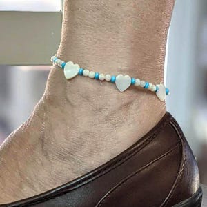 May include: Anklet featuring white heart-shaped charms and small blue and white beads. The delicate jewelry is worn on the ankle, paired with a brown shoe.