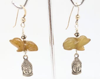 May include: A pair of dangle earrings featuring a carved jade green pig charm and a silver Buddha pendant. The earrings are made with silver and gold accents.