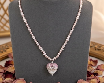 Puede incluir: Un collar que presenta una hilera de pequeñas perlas de agua dulce de color rosa claro. La pieza central es un colgante en forma de corazón con un degradado de rosa suave y blanco, decorado con intrincados patrones de filigrana plateada.