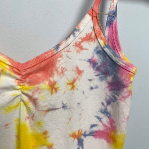 May include: A white tank top with a tie-dye pattern in shades of pink, yellow, and blue. The straps are thin and adjustable.