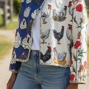 May include: A quilted jacket with a split design. One side has a blue background with white and black chicken illustrations, the other a white background with chicken and floral patterns. The jacket is worn with blue jeans.
