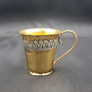 May include: A gold-coloured metal cup with a silver-coloured design around the rim. The cup has a handle and a rounded bottom.