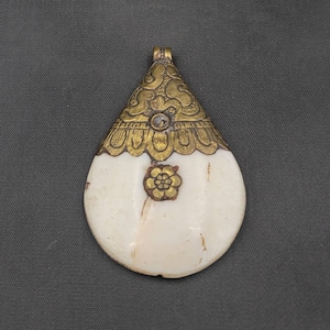 May include: A white bone pendant with gold accents. The pendant is shaped like a teardrop and has a floral design on the front. The pendant is attached to a gold chain.