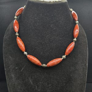 Könnte beinhalten: Eine Halskette mit abwechselnd länglichen, spindelförmigen orange-roten Perlen und kleinen, runden, metallisch glänzenden Zwischenperlen. Die Perlen sind in einem sich wiederholenden Muster entlang des Strangs angeordnet.