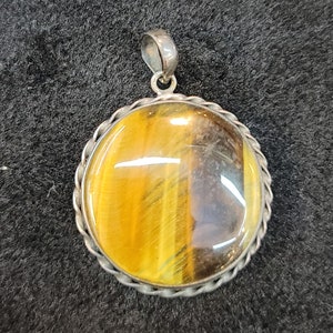 Może przedstawiać: Srebrny wisiorek z okrągłym, kaboszonowym oczkiem tygrysim. Kamień ma złotobrązowy kolor z ciemnobrązowymi paskami.