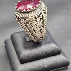 May include: A silver ring with an ornate design featuring a large, oval-shaped ruby gemstone.