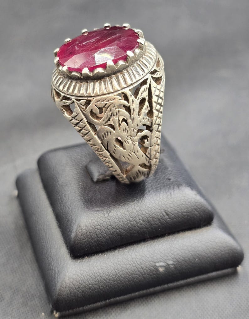 May include: A silver ring with an ornate design featuring a large, oval-shaped ruby gemstone.