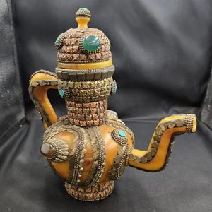 Op de afbeelding: Een sierlijke, amberkleurige theepot met ingewikkeld metaalwerk en turquoise en groene stenen accenten. De theepot heeft een decoratief deksel en tuit, met gedetailleerde patronen en versieringen. Een uniek en artistiek stuk.