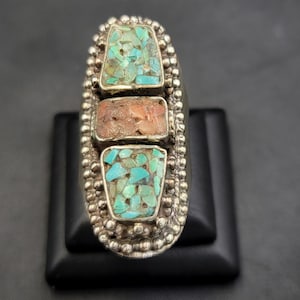 Könnte beinhalten: Ein silberner Ring mit drei rechteckigen Steinen, die darin eingefasst sind. Der obere Stein ist türkis, der mittlere Stein ist rosa und der untere Stein ist türkis. Der Ring ist mit kleinen silbernen Perlen verziert.