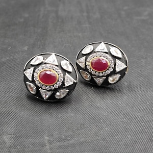May include: A pair of oval-shaped stud earrings featuring a central oval red stone surrounded by a halo of small clear stones. The dark metal base is accented with alternating triangular and teardrop-shaped clear stones arranged in a radial pattern.