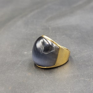 Puede incluir: Un anillo de oro con una piedra ovalada negra. La piedra tiene un patrón oscuro y arremolinado.