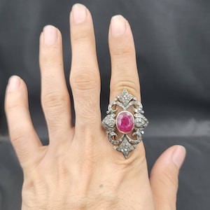 May include: A silver ring featuring a large, oval ruby gemstone surrounded by smaller diamonds. The ring has an intricate, ornate design.
