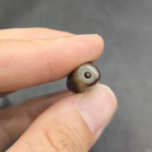Puede incluir: Una cuenta peque&ntilde;a, lisa y de forma ovalada con una superficie oscura de color marr&oacute;n y gris moteado. La cuenta presenta un &uacute;nico orificio circular centrado perforado en su extremo, revelando un interior oscuro. Tiene un acabado pulido.