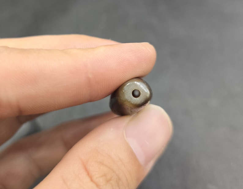 Puede incluir: Una cuenta peque&ntilde;a, lisa y de forma ovalada con una superficie oscura de color marr&oacute;n y gris moteado. La cuenta presenta un &uacute;nico orificio circular centrado perforado en su extremo, revelando un interior oscuro. Tiene un acabado pulido.