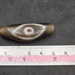 Puede incluir: Una cuenta de piedra marr&oacute;n, alargada y cil&iacute;ndrica, con un patr&oacute;n blanco en forma de ojo y un centro oscuro. La cuenta est&aacute; colocada sobre una regla met&aacute;lica, mostrando una longitud de aproximadamente 6,3 cm, con textura y desgaste visibles.