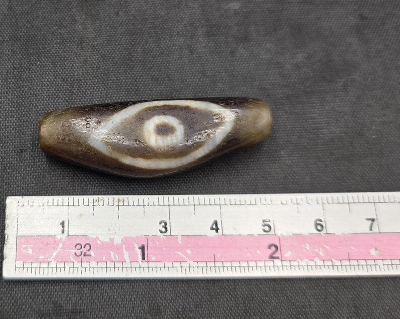 Puede incluir: Una cuenta de piedra marr&oacute;n, alargada y cil&iacute;ndrica, con un patr&oacute;n blanco en forma de ojo y un centro oscuro. La cuenta est&aacute; colocada sobre una regla met&aacute;lica, mostrando una longitud de aproximadamente 6,3 cm, con textura y desgaste visibles.