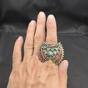 Könnte beinhalten: Ein großer, verzierter Silberring mit schildförmigem Design. Der Ring hat türkisfarbene und rote Steinakzente mit einem zentralen Türkisstein. Der Ring wird an einer Hand vor dunklem Hintergrund getragen.