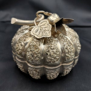 May include: A silver pumpkin-shaped trinket box with intricate floral designs and a detailed stem with a leaf and flower.