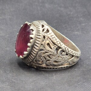 May include: A silver ring with an ornate design and a large, oval-shaped, dark pink gemstone.