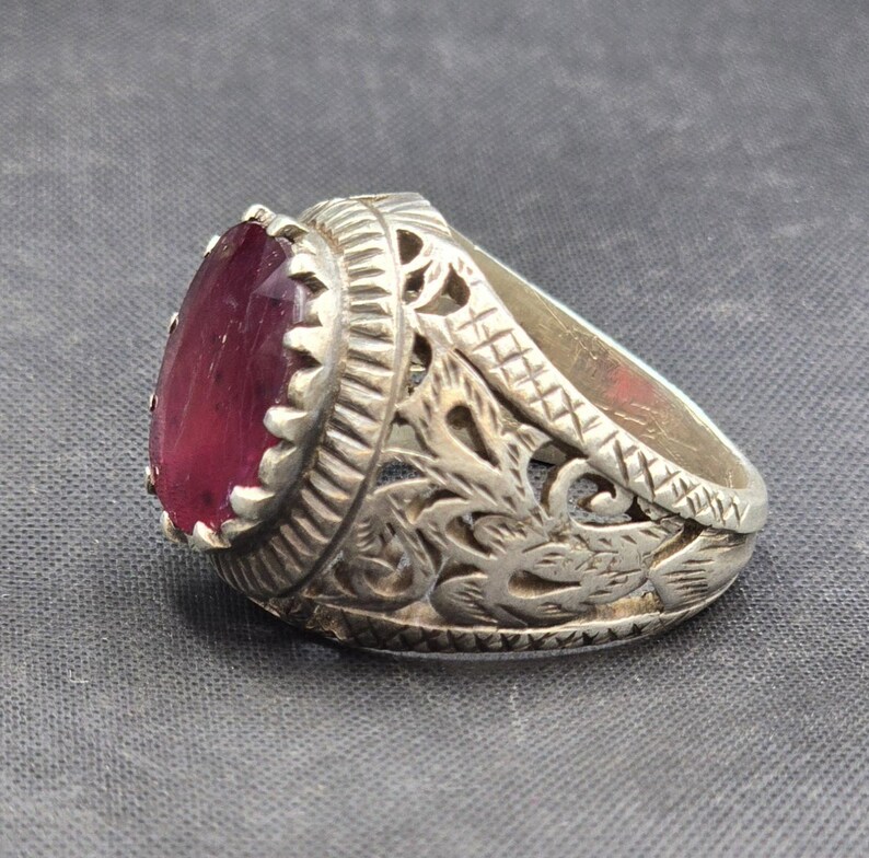 May include: A silver ring with an ornate design and a large, oval-shaped, dark pink gemstone.
