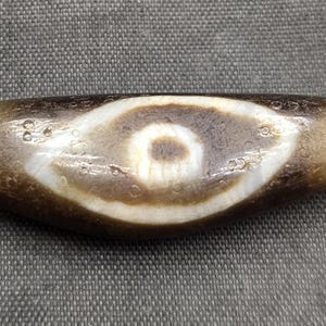Puede incluir: Una cuenta de piedra cil&iacute;ndrica y alargada con una superficie de color marr&oacute;n oscuro desgastada y un patr&oacute;n prominente en forma de ojo en blanco y marr&oacute;n oscuro en el centro. La cuenta tiene una textura suave y pulida con porosidad natural.