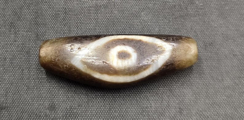 Puede incluir: Una cuenta de piedra cil&iacute;ndrica y alargada con una superficie de color marr&oacute;n oscuro desgastada y un patr&oacute;n prominente en forma de ojo en blanco y marr&oacute;n oscuro en el centro. La cuenta tiene una textura suave y pulida con porosidad natural.