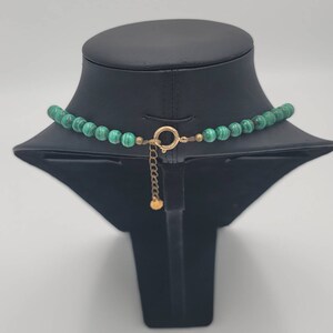 May include: A green malachite bead necklace with a gold clasp and chain. The necklace is displayed on a black mannequin bust.
