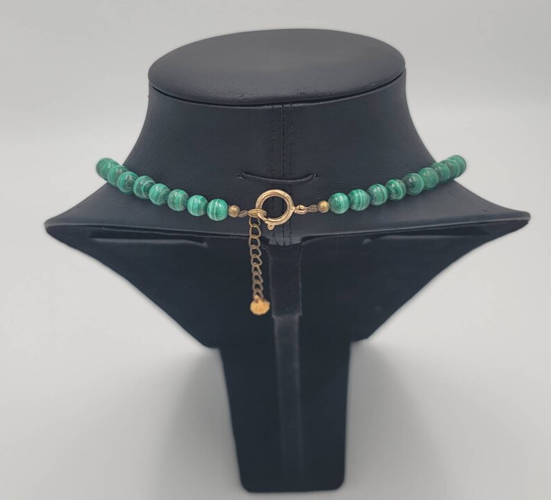 May include: A green malachite bead necklace with a gold clasp and chain. The necklace is displayed on a black mannequin bust.