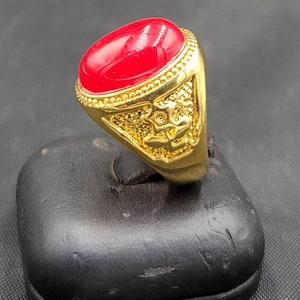 Op de afbeelding: Een goudkleurige ring met een grote, ovale, rode edelsteen. De ring heeft ingewikkelde details rond de band en de zetting. De ring wordt gepresenteerd op een zwarte standaard.