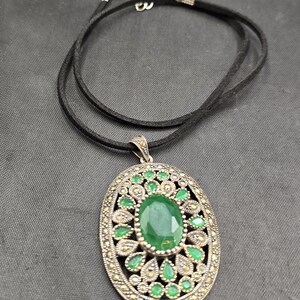 May include: Oval pendant necklace with a large green gemstone center and smaller green stones around the edge. The pendant is set in a silver-colored metal with a black cord necklace.