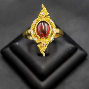 May include: A gold ring with a red gemstone set in a detailed, ornate design. The ring features intricate carvings and small pink gemstones.