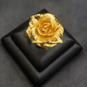 May include: A gold-colored rose ring with a detailed design. The ring is displayed on a black velvet cushion.