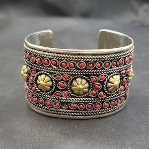 Könnte beinhalten: Silbernes Armreif-Armband mit aufwendigen Details. Das Armband verfügt über ein Band aus roten Perlen und goldfarbenen floralen Akzenten.