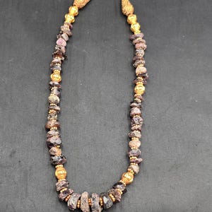 May include: A beaded necklace featuring dark purple and brown gemstone beads interspersed with gold-colored metal beads. The necklace has a gold-colored clasp and decorative elements at the top. The gemstones vary in size and shape.