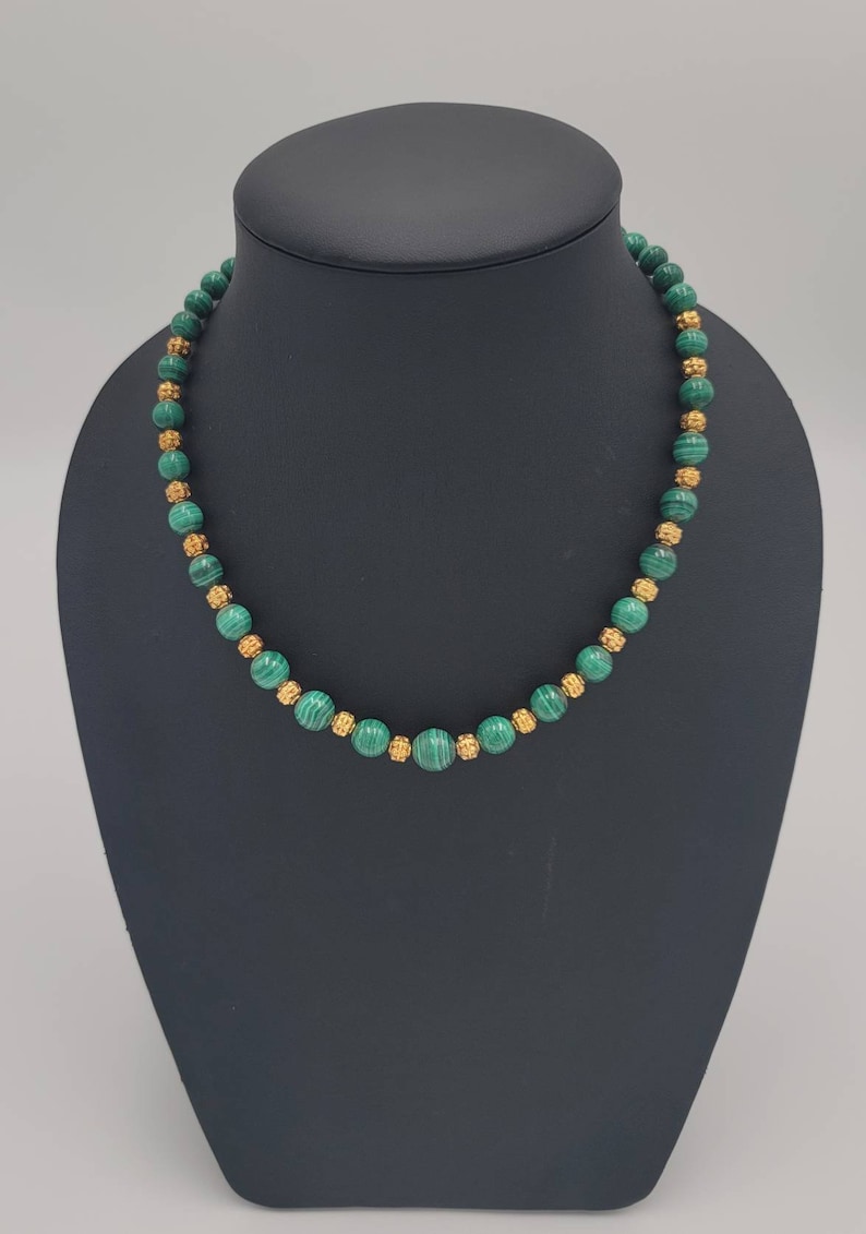 May include: A green malachite and gold bead necklace on a black display stand. The necklace is made of round beads with a gold finish.