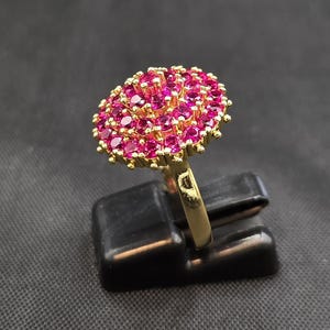 Puede incluir: Un anillo dorado con un gran grupo ovalado de pequeñas piedras preciosas de color rosa brillante. Las piedras están densamente agrupadas. El anillo se presenta sobre un soporte negro, sobre un fondo gris oscuro.