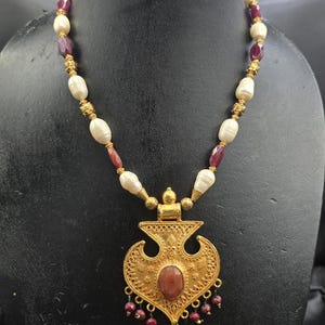 May include: A gold-toned necklace featuring a large pendant with a central oval stone and dangling red beads. The necklace has alternating cream-colored pearls and red gemstones, with gold-colored accents.