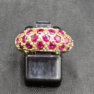 Puede incluir: Un anillo dorado con un grupo de gemas redondas de color rubí vibrante. El engaste del anillo presenta un diseño texturizado, y las gemas están muy juntas, creando un efecto deslumbrante. El anillo se exhibe sobre un soporte negro.