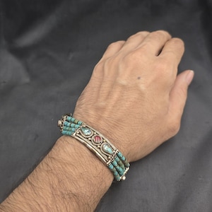 May include: A turquoise beaded bracelet with a silver metal centerpiece. The centerpiece features three oval-shaped stones in turquoise and red. The bracelet is worn on a wrist.