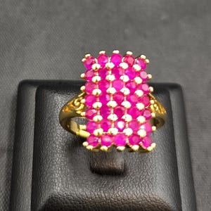 May include: A gold-toned ring featuring a cluster of vibrant pink gemstones. The ring's design showcases a floral arrangement of the pink stones, set against a detailed gold band. The ring is displayed on a black stand.