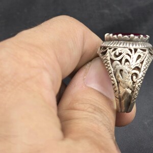 May include: A silver ring with an ornate design featuring a red gemstone. The ring has a detailed filigree pattern and a raised setting for the stone.