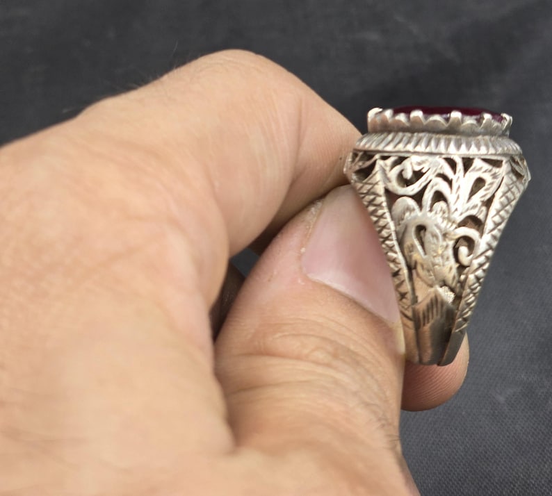 May include: A silver ring with an ornate design featuring a red gemstone. The ring has a detailed filigree pattern and a raised setting for the stone.