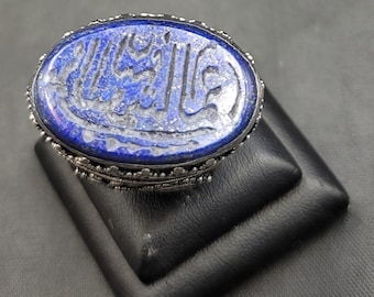 Può includere: Un anello d'argento con una pietra ovale in lapislazzuli blu. La pietra è incisa con calligrafia araba intricata.