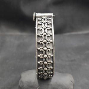Puede incluir: Un brazalete de plata con un diseño decorativo de formas de media luna repetidas y pequeños puntos. El brazalete tiene un cierre con bisagra.