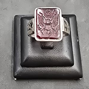 Könnte beinhalten: Ein silberner Ring mit einem dunkelroten Stein, der ein eingraviertes Bild eines Adlers mit einem Schild auf der Brust zeigt. Der Ring wird auf einem schwarzen Ringständer präsentiert.